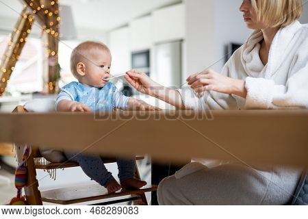Mother Wearing Cosy Bathrope Spoon Feeding Her Baby Boy Child In Baby High Chair With Fruit Puree At