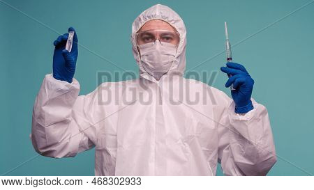 A Male Doctor In Protective Overalls And A Mask Holds An Ampoule With A Vaccine And Syringe In His H