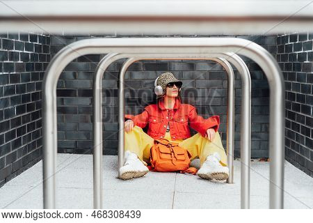 Hipster Fashion Young Woman In Bright Clothes, Sun Glasses, Bucket Hat And Earphones Sitting Near Gr