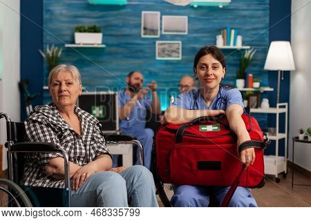 Female Healthcare Assistant Sitting In Living Room Of Nursing Home Next To Elderly Woman In Wheelcha