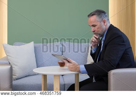 Senior Businessman In Suit And Necktie Reading Email On Tablet Computer In Cafeteria During Coffee B