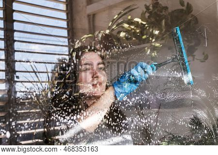 A Girl Washes Windows At Home. Woman Cleaning Window With Small Squeegee. Reflections Of The Balcony