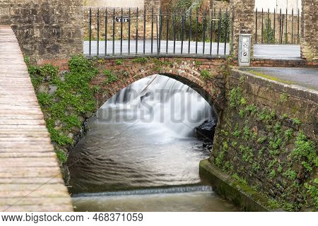 Long Exposure Of A Watefall Flowing Under A Bridge On The River Lim Walkway At Lyme Regis In Dorset.