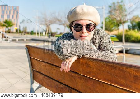 Portrait Of A Young Caucasian Woman Looking At The Camera, Resting On A Garden Bench In A Park On An