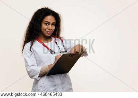 Friendly Dark Skinned Female Doctor In Uniform With Stethoscope Filling Medical Form On Light Backgr