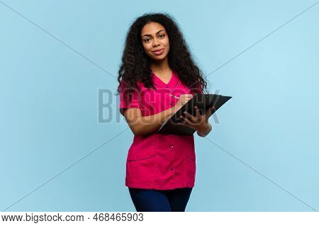 Friendly Dark Skinned Female Nurse In Pink Uniform Filling Medical Form On Blue Background. African 