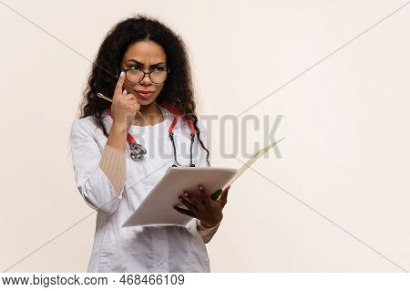 Emotional Puzzled Dark Skinned Female Doctor In Uniform With Glasses And Stethoscope On Light Backgr