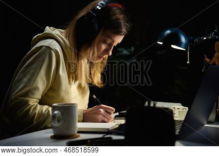 Female Sudent Wearing Headphones Studying At Night Room, Using Laptop. Online Education At Home.