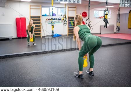 Young Woman With Blonde Hair Squatting With Kettlebells In Gym