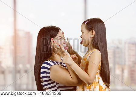 Side View Of Woman And Little Girl Are Making Faces To Each Other. Blurred Windows Background.