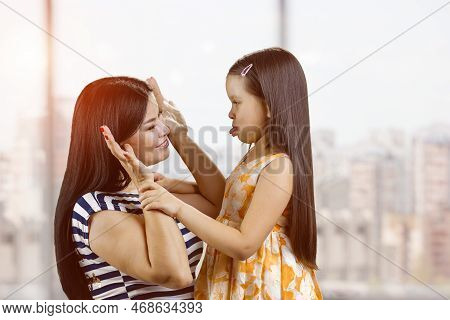 Young Mother And Her Daughter Are Fooling Around Making Faces. Little Girl Is Showing Tongue To Her 