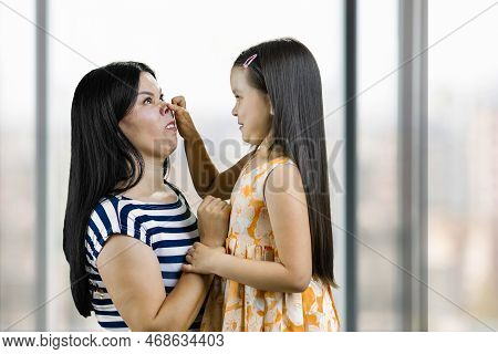 Little Girl Is Making Pig Nose To Her Mother. Having Fun And Fooling Around. Indoor Blurred Window B