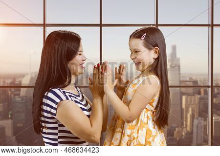 Side View Of Woman And Her Daughter Are Playing Clapping Game. Checkered Window Background With City