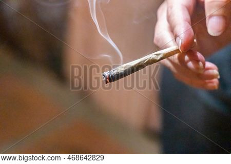 Finger Holding Lighted Cannabis Cigarette With Smoke In Dark Room, With Selective Focus On Cannibis 