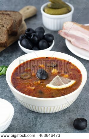 Traditional Russian Soup Solyanka With Meat, Sausages, Pickles, Olives, Rye Bread And Sour Cream.