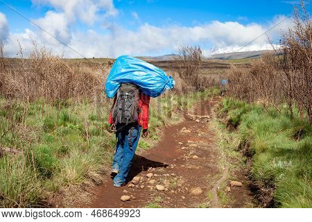 A Porter Carrying Heavy Load On His Back Walks Along The Road.