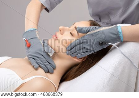 A Cosmetologist Doctor Makes A Microcurrent Massage Procedure On The Face And Neck Of A Young Beauti