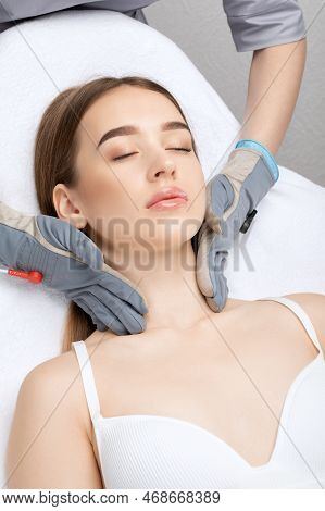 A Cosmetologist Doctor Makes A Microcurrent Massage Procedure On The Face And Neck Of A Young Beauti