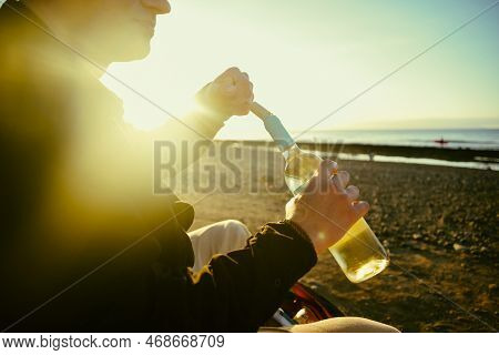 A Man On The Shore Of The Sea Or Ocean Opens A Bottle Of White Wine. A Date On The Beach, Alcohol On