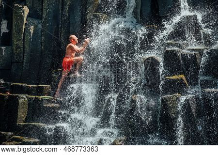 Caucasian Man In Swimsuit Washing His Face Under The Falling Water Streams Flowing On Black Volcanic