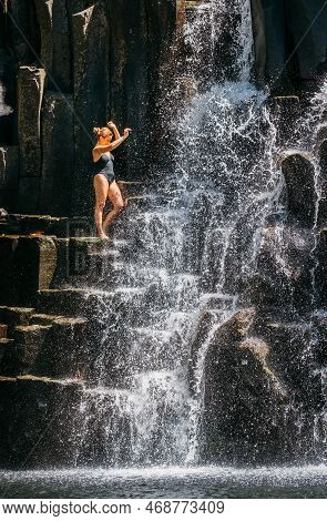 Caucasian Woman In Black Swimsuit Refreshing Under The Falling Water Streams Flowing On Black Volcan