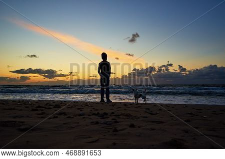 Boy Silhouette And Dog Standing On Beach Near Sea And Looking On The Waves In Sunset