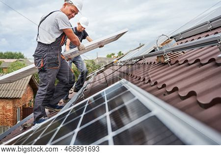 Men Technicians Carrying Photovoltaic Solar Moduls On Roof Of House. Builders In Helmets Installing 