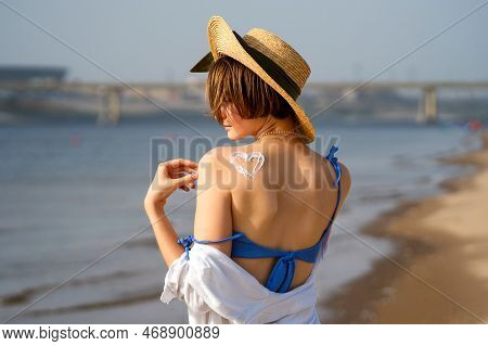Young Woman In Straw Sunhat With Sun Cream On Tanned Back In Form Heartlet, Shape Heart. Skincare. C
