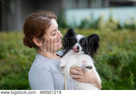 Caucasian Red-haired Woman Holding Pappilion Dog In Her Arms Outdoors. Black And White Continental S