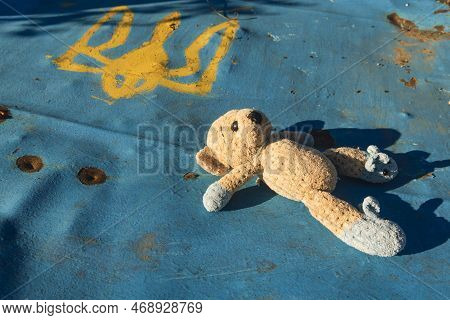 A Toy (teddy Bear) Lies On A Damaged Fence With The State Emblem Of Ukraine. War In Ukraine. Russian