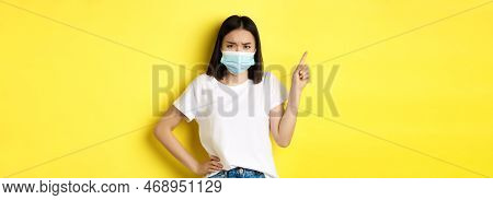 Covid, Health Care And Pandemic Concept. Asian Female Model In Medical Mask And White T-shirt Pointi