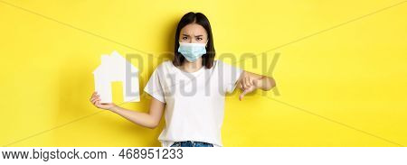 Covid-19 And Real Estate Concept. Disappointed Asian Woman In Medical Mask, Showing Thumb Down And P