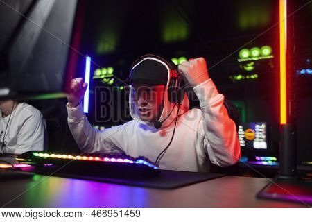 Young Emotional Guy Gamer In Headphones Sits In A Computer Club And Plays A Computer Game, A Man Scr