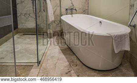 Large Oval Acrylic Bathtub In The Bathroom. A White Towel Is Hung On The Edge. On The Tiled Beige Wa
