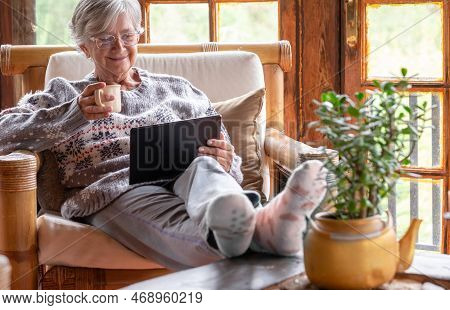Smiling Relaxed Pensioner Woman Sitting At Home In An Armchair Using A Digital Tablet To See Videos 