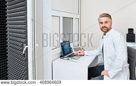 Portrait Of Male Audiologist Sitting In His Office Of Audiology Center Near Audiometric Booth For Au