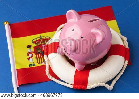 Piggy Bank, Lifebuoy And Flag On A Blue Background, Concept On The Theme Of Saving The Spanish Econo