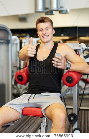 sport, bodybuilding, lifestyle, technology and people concept - smiling young man with smartphone showing thumbs up gesture in gym