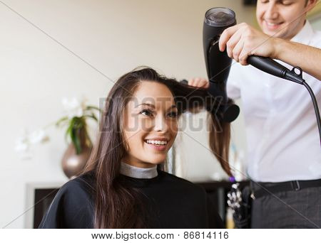 beauty, hairstyle and people concept - happy young woman and hairdresser with fan making hot styling at hair salon