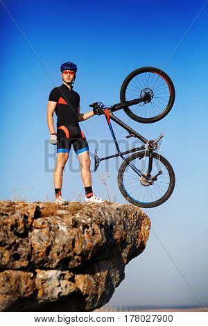 Man biker holding bike on the top of a hill against beautiful blue sky. Extremal sport concept. Beautiful landscape. Power and energy. Sportsman in the blacksportwear, helmet and sunglasses. Travel in the countryside. Spring season.