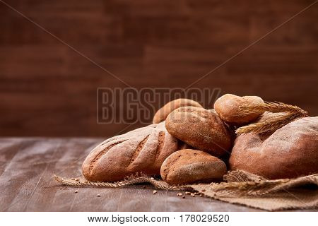 handmade tasty bread lying on burlap on the wooden table with flour, wheat and ears of wheat. Brown blurred background. Brown bread and rulls. Delicious food. Fresh baking. Tasty and appetizing.