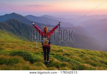 Redheaded girl athlete with a backpack and sticks stands on the green hillocks and looks at high mountain landscapes and fascinating sunset.