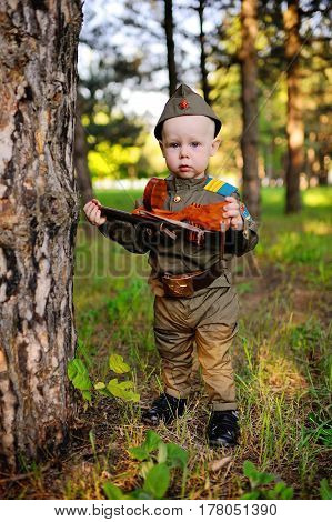 A cute child in the uniform of a Soviet soldier with a St. George ribbon with a toy gun on the background of nature. May 9, February 23, defender, against fascism.