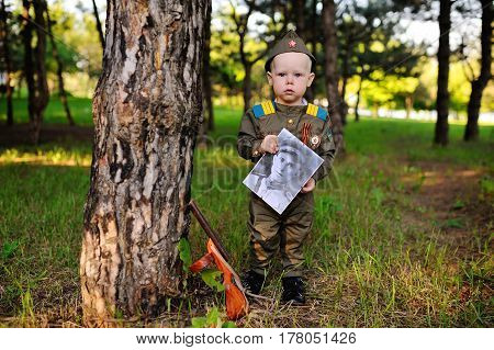 Child boy in military uniform of a Soviet soldier with a photograph of a veteran of World War II in hands against a background of green trees. Victory Day, May 9, against fascism.