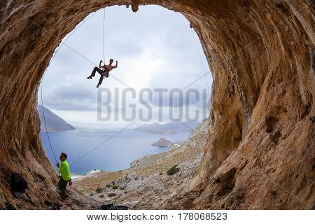 Rock climbers in cave: leading climber flexing muscles jokingly his partner belaying