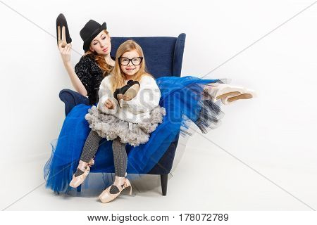 The elder sister and little girl ballerina are sitting in a chair. They keep their shoes on high heels. Girls in ballet tutu and pointe shoes. Lovely girl. Family tradition