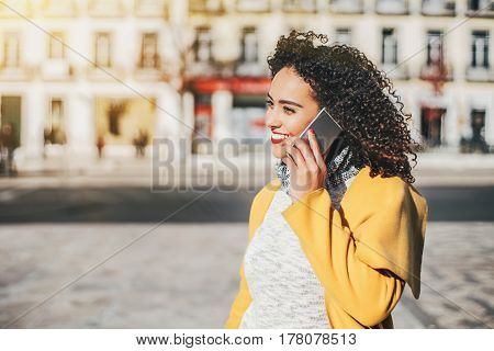 Beautiful Brazilian curly woman talking by phone with friend while standing on sunny street stylish hipster brunette lady using smartphone for call her sister with copy space for your text message