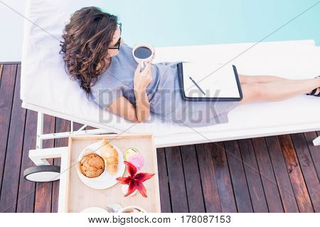 Young woman having cup of tea and relaxing on a sun lounger near poolside