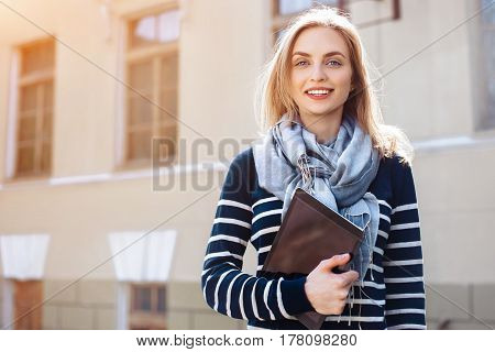 Young cheerful female student is enjoying free timewhile is wallking with touch pad in outdoors.Beautiful woman is holding digital tablet and smiling for someone during rest in outdoors