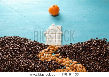 The house from white chocolate, the earth from coffee grains, the road from almonds, the sun from a citrus on blue background. Abstract symbols flat lay composition. Concept of realty, cosiness, comfort, happy family etc.
